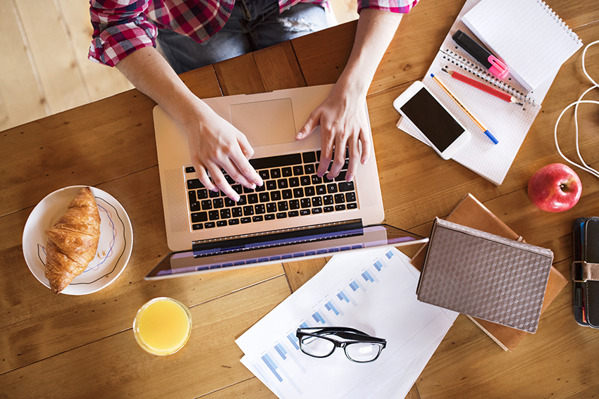 Unrecognizable teenage girl in checked shirt at home sitting at the table, working on laptop, studying.